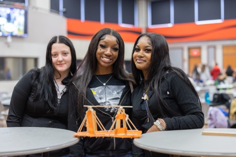 3 students smiling with their bridge, suspended between two tables to be weighed down for a stress test
